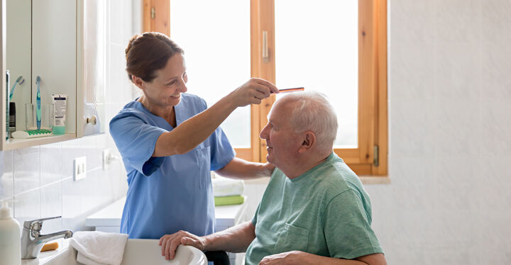 Eine Pflegerin kämmt einem vor einem Waschbecken sitzenden älteren Mann die Haare.