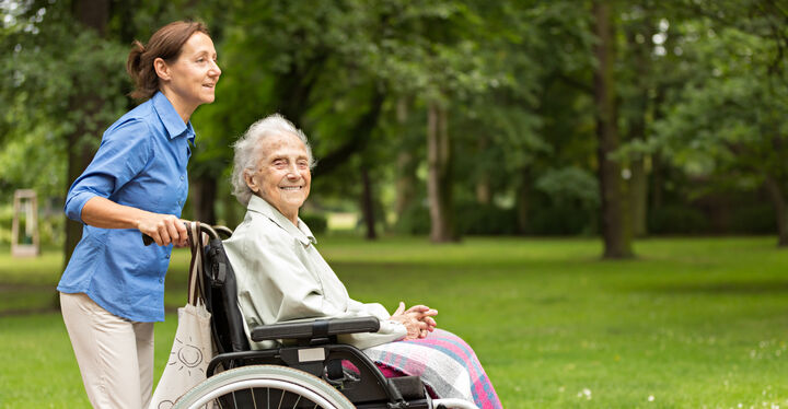 Eine Frau rollt eine in einem Rollstuhl sitzende ältere Frau durch den Park und die ältere Frau lächelt dabei in die Kamera.